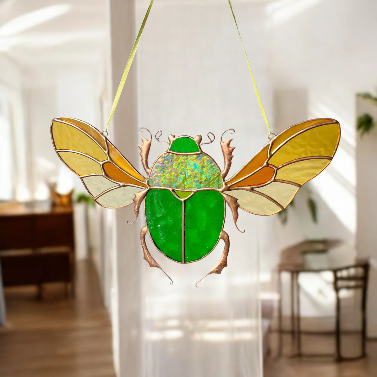 A stained-glass hanging beetle ornament with a bright green body, iridescent green-blue wing area, and tan wings outlined in gold-toned metal, suspended by thin yellow ribbons in a softly blurred indoor room.