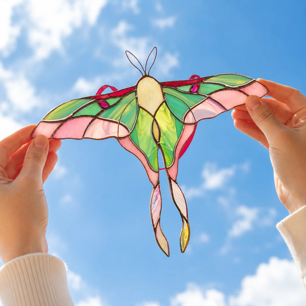 A stained-glass moth ornament with pink, green, and cream panels is held up by two hands against a blue sky with clouds.