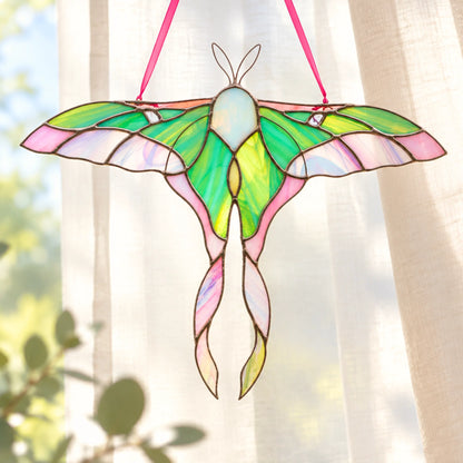 A stained glass pink moth suncatcher with vibrant green, pink, and yellow wings, suspended by a pink ribbon in front of a soft-focus window with sheer curtains.