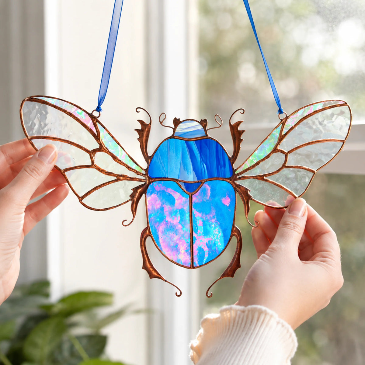 A stained-glass beetle window hanging ornament with iridescent blue and pink sections, brown metal outlines, and clear wing panels, held by two hands in front of a bright window.