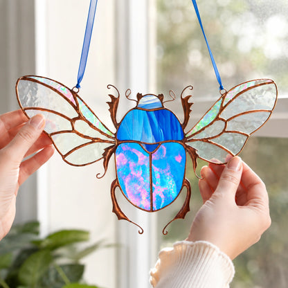A stained-glass beetle window hanging ornament with iridescent blue and pink sections, brown metal outlines, and clear wing panels, held by two hands in front of a bright window.