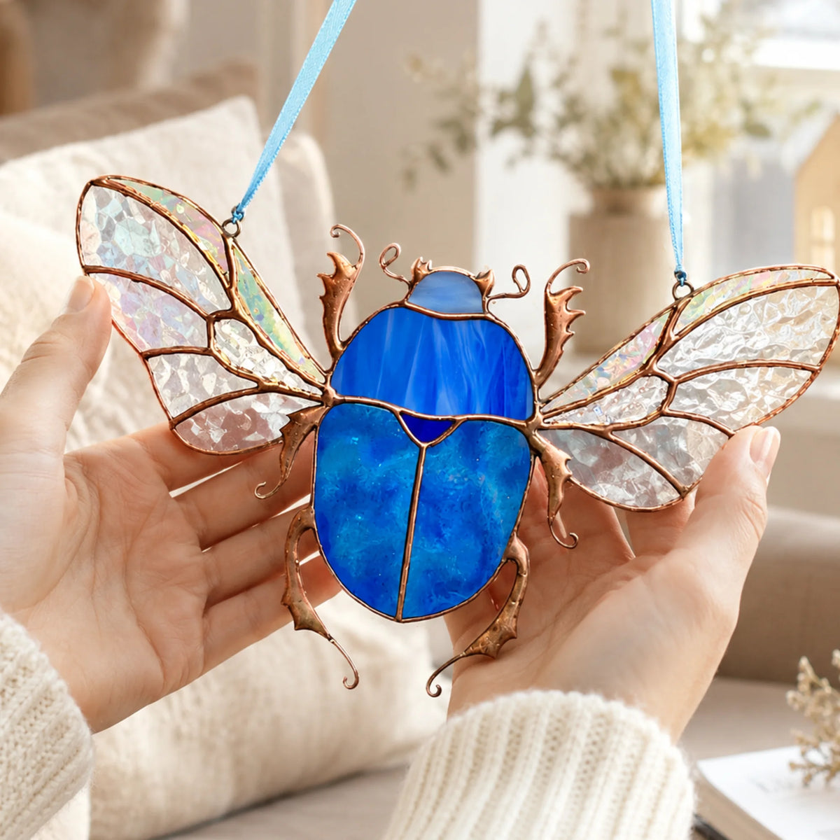 A stained-glass beetle ornament with a bright blue body and iridescent white wings, held in two hands against a softly blurred indoor background.