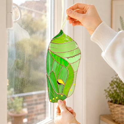 A green stained-glass luna moth chrysalis ornament with yellow and black details is held up by two hands in front of a bright window.