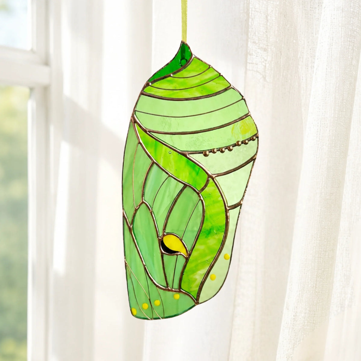 A gorgeous green stained glass butterfly chrysalis hanging against a soft, blurred background, serving as an enchanting decorative gift.