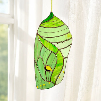 A gorgeous green stained glass butterfly chrysalis hanging against a soft, blurred background, serving as an enchanting decorative gift.