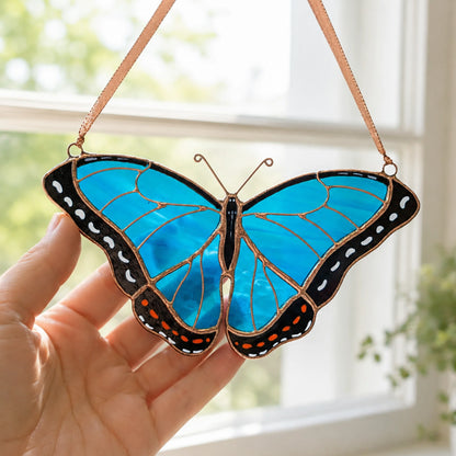 A stained glass morpho butterfly ornament with vibrant blue wings, black borders, and orange accents, held by a hand in front of a bright window.