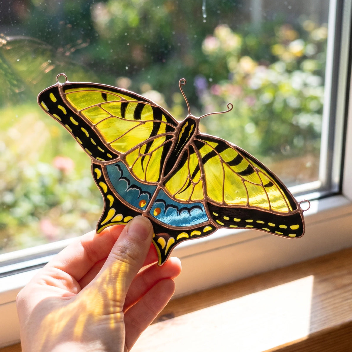 A hand holds a stained glass swallowtail butterfly suncatcher with yellow, black, and blue glass panels in front of a sunlit window.