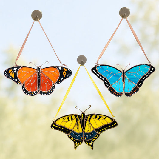 Three butterfly hanging ornaments with colorful wings are displayed against a soft, blurred outdoor background. The ornaments feature monarch, morpho and swallowtail butterflies