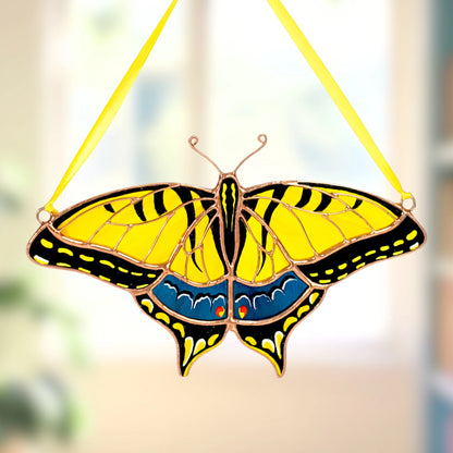 A stained glass swallowtail butterfly ornament with yellow, black, and blue wings, suspended by a yellow ribbon against a softly blurred indoor background.
