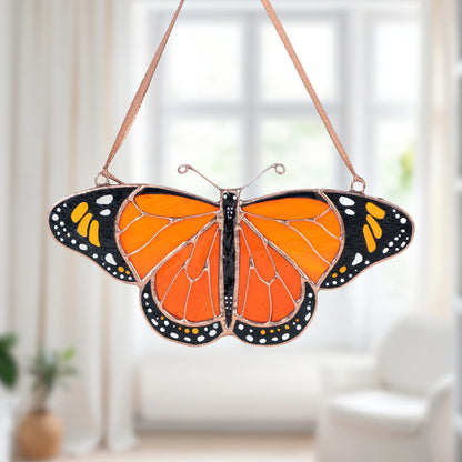 A stained glass butterfly ornament with vibrant orange, black, and white sections, suspended by thin brown strings against a softly blurred bright room background.