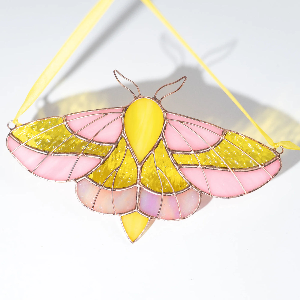 A stained glass moth ornament with pink and yellow art glass, held together by copper-colored wire and suspended by a yellow ribbon, shown on a white background.