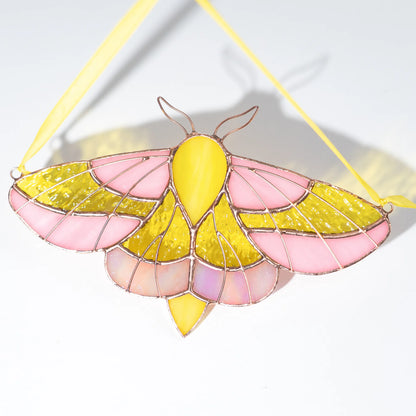 A stained glass moth ornament with pink and yellow art glass, held together by copper-colored wire and suspended by a yellow ribbon, shown on a white background.