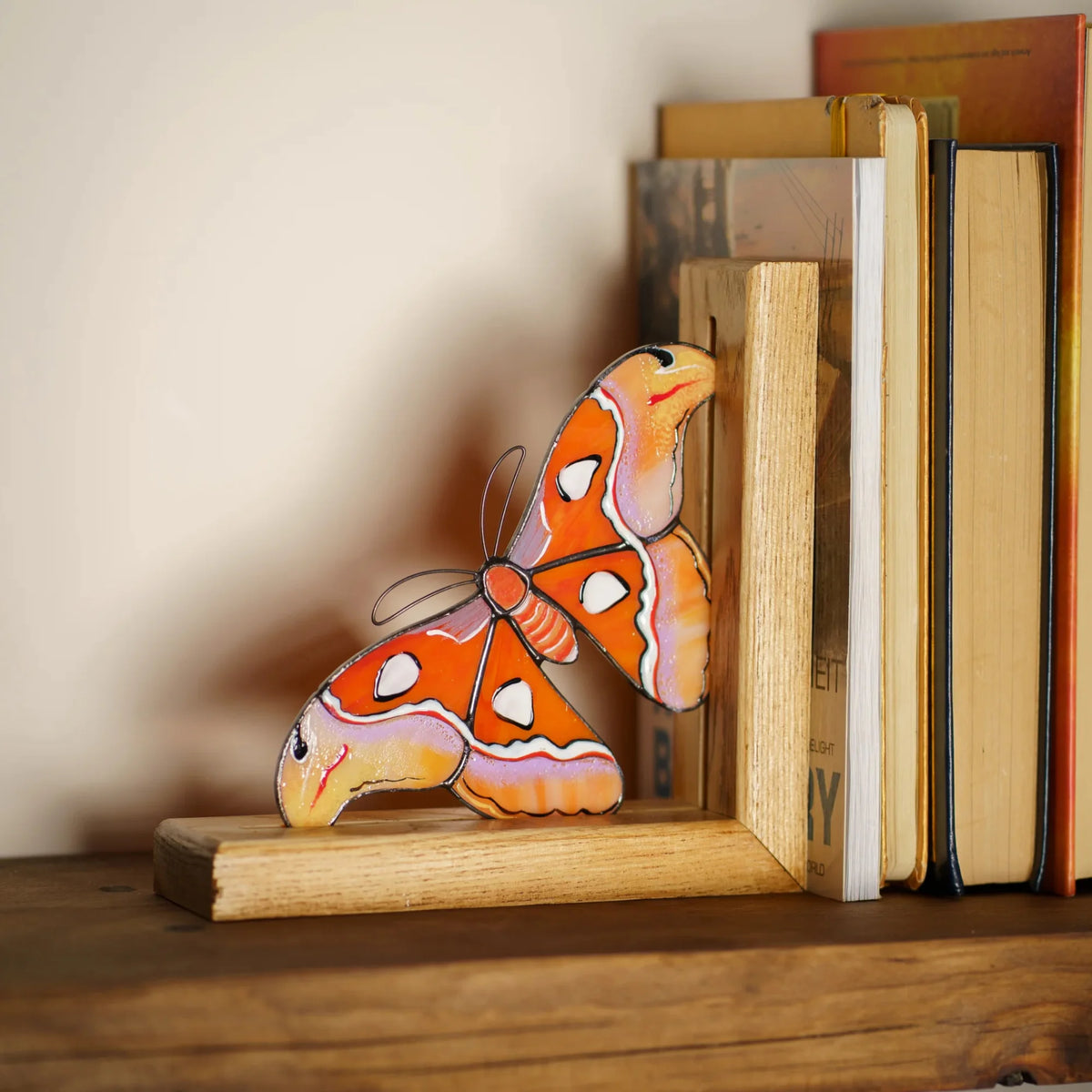 An orange and yellow stained glass moth bookend elegantly supporting a row of books on a wooden shelf, serving as a charming home library accent.