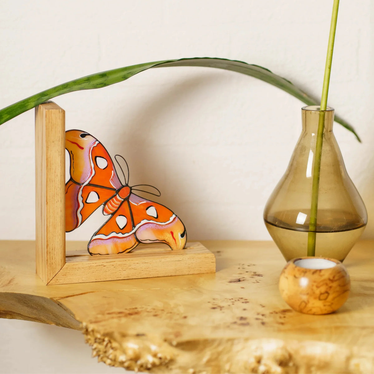 An orange and yellow stained glass moth bookend styled on a wooden shelf next to a decorative glass vase and plant stem, creating an exquisite display.