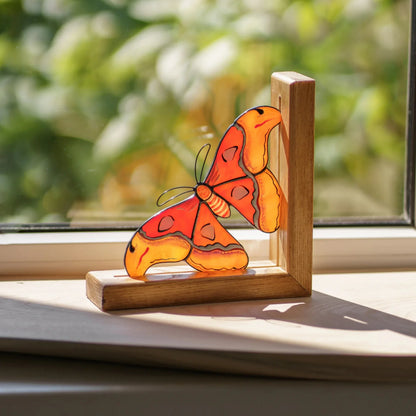 A vibrant orange and yellow stained glass moth bookend captured on a sunny windowsill, highlighting its exceptional craftsmanship and warm glow.