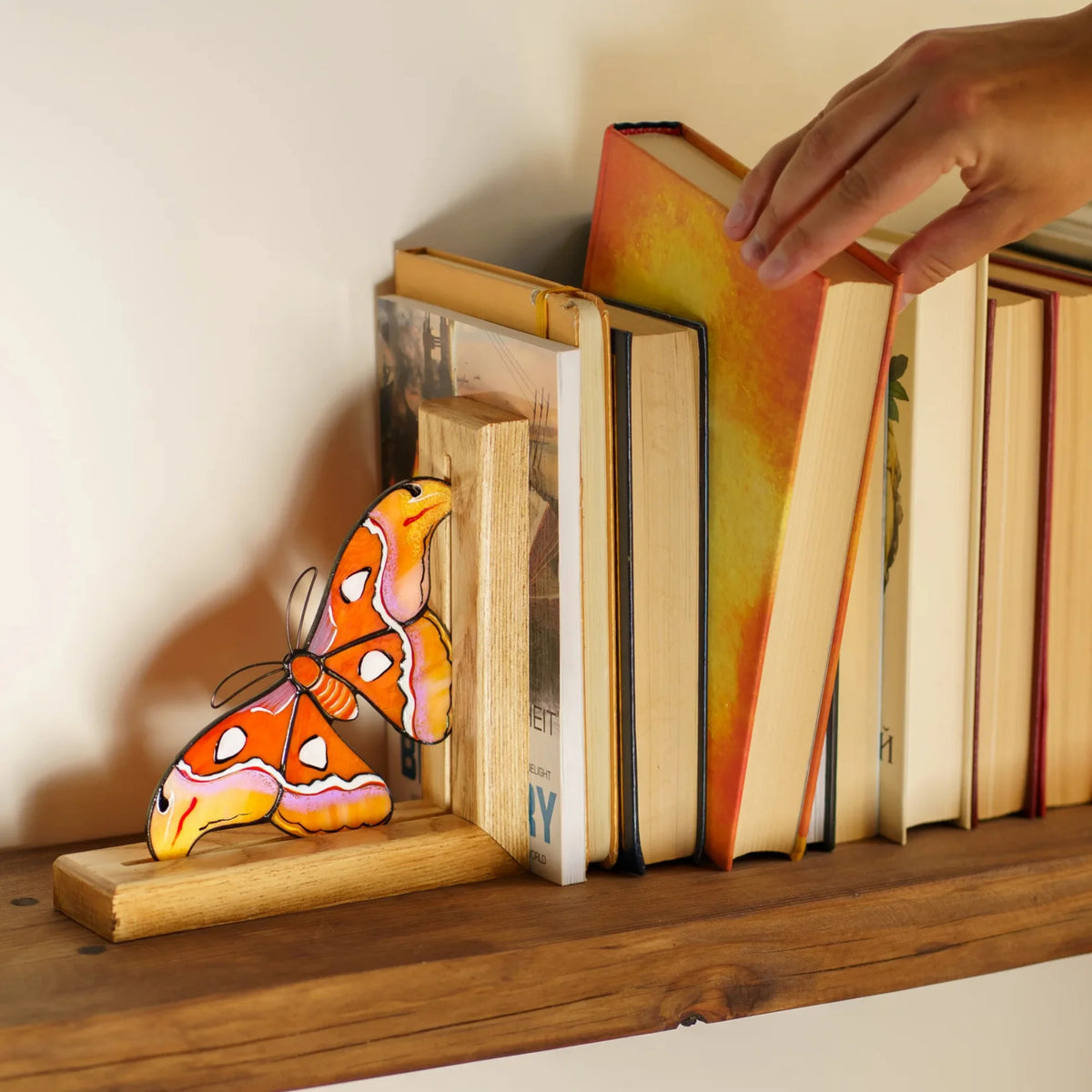 A person gently adjusting a book next to an orange and yellow stained glass moth bookend on a shelf, showcasing its delightful utility and appeal as a gift.