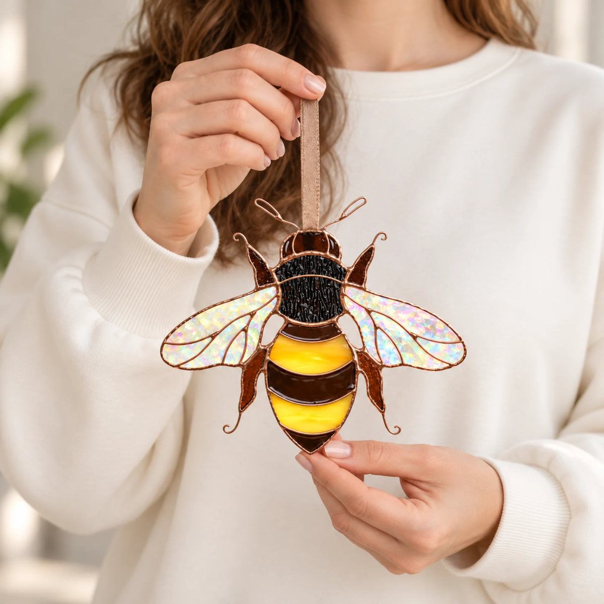 Woman in a cream sweater holding the small glass bee by its ribbon to show its size