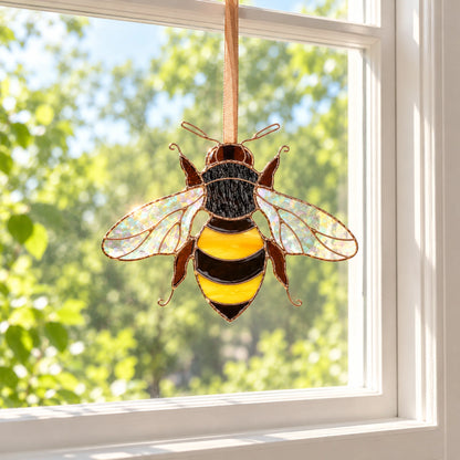 Small bee suncatcher hanging on a ribbon in the center of a white-framed window with trees outside