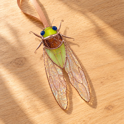 Green stained glass cicada lying on a wooden table in sunlight, showing copper legs and iridescent stained glass cicada wings