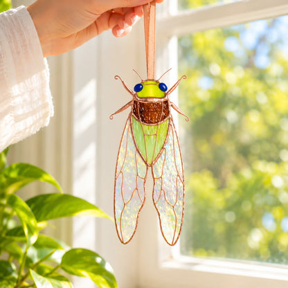Hand hanging a green stained glass cicada suncatcher in a bright window beside a houseplant, stained glass insect window decor