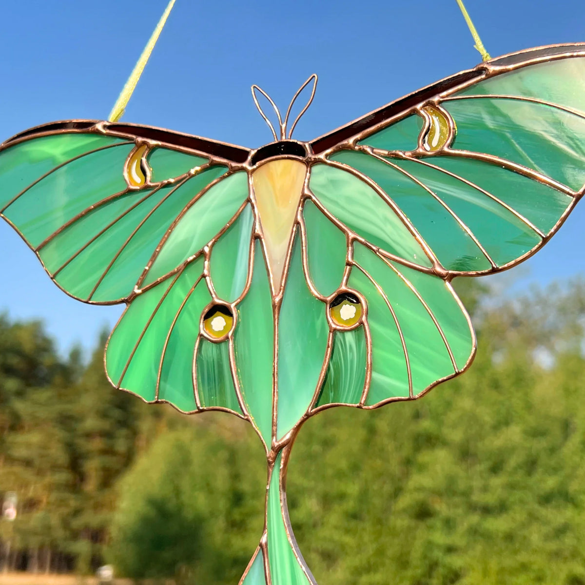 A close-up view of stained glass luna moth suncatcher with vibrant green and teal wings, gold metal outlines, and small gold eye details, shown hanging outdoors against a blue sky and blurred green trees.