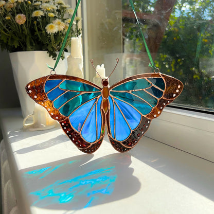 A stained glass butterfly suncatcher with blue wings and copper-gold borders, hanging by green strings on a sunny windowsill with a flower pot in the background.