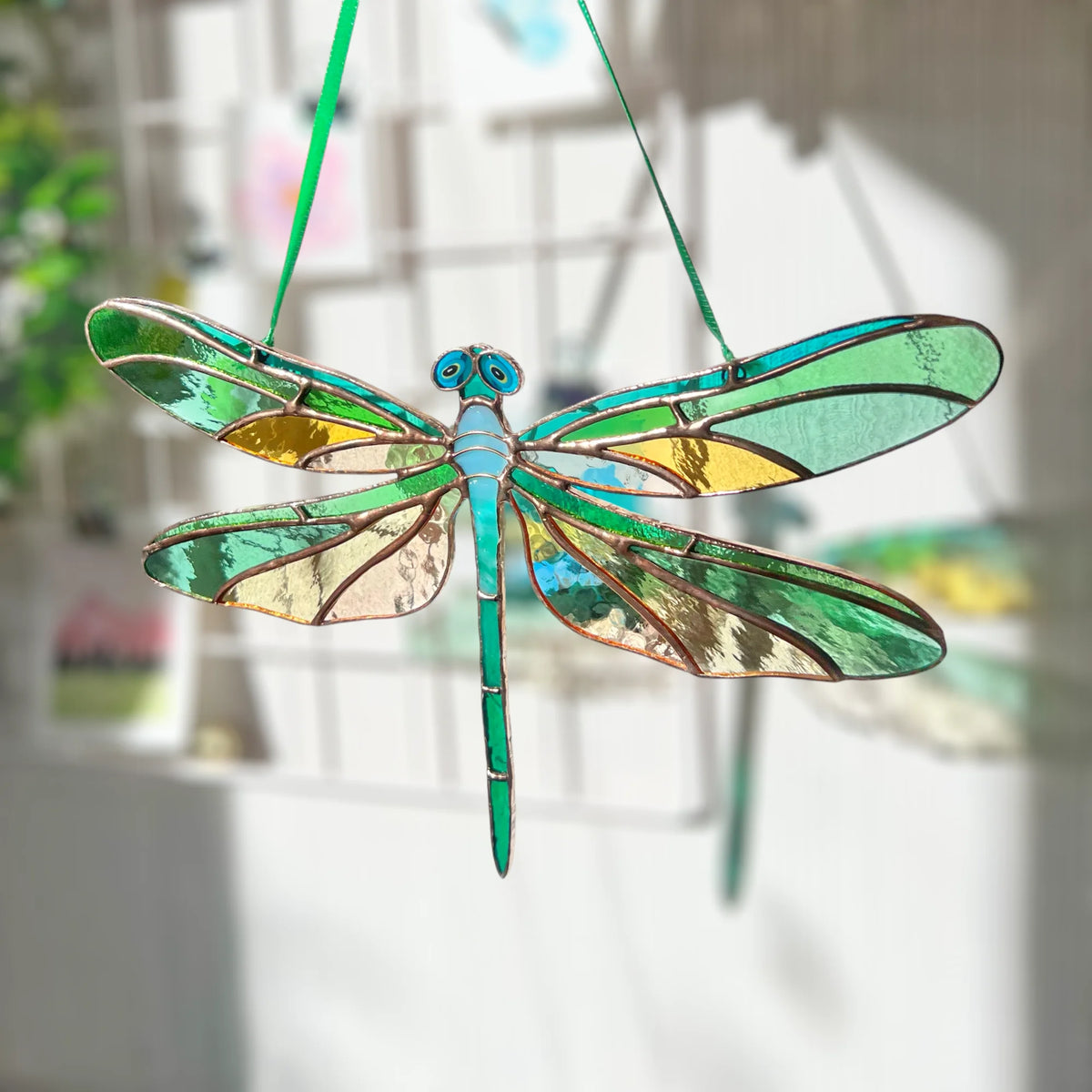 A stained glass dragonfly ornament hanging by a green string, featuring green, blue, and gold glass panels outlined in copper-colored metal, set against a softly blurred indoor background.