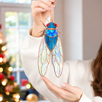 Woman holding a blue stained glass cicada by its ribbon beside a lit Christmas tree, a handmade stained glass insect ornament and gift idea