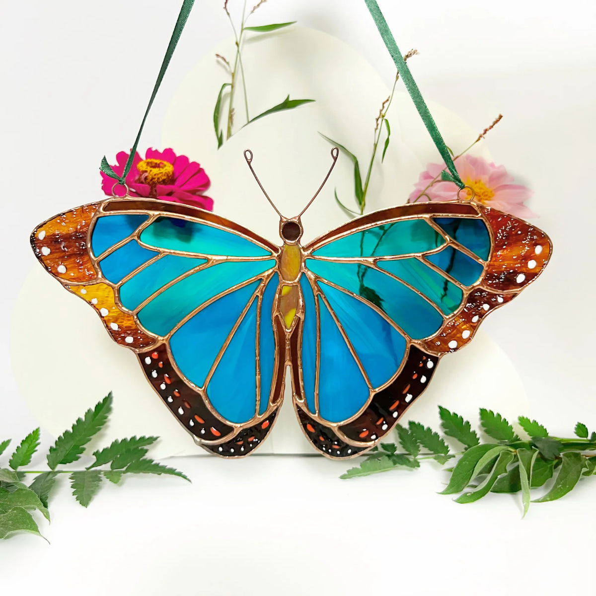 A stained glass butterfly ornament with vibrant blue wings, brown and orange patterned borders, and green hanging ribbons, displayed against a white background with decorative flowers and leaves.