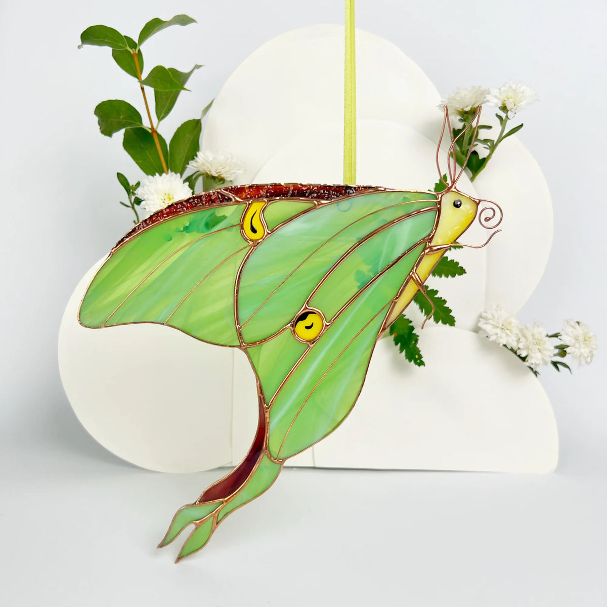 A stained glass luna moth ornament hanging, decorated with green glass wings, yellow eyes, and copper wire outlines, with small white flowers and green leaves around it.