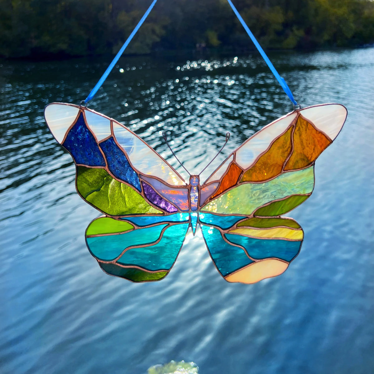 A stained glass butterfly hanging outdoors by blue strings, with a blurred lake and trees in the background. The butterfly features multiple colors including blue, green, yellow, orange, and purple.
