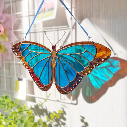 A stained glass morpho butterfly suncatcher with vibrant blue wings, brown borders, and red accents, suspended by a blue ribbon against a bright window background.