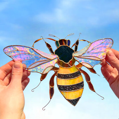A stained glass bumblebee ornament with iridescent purple and blue wings, a black and yellow striped body, and gold-toned metal framing, held by two hands against a blue sky background.
