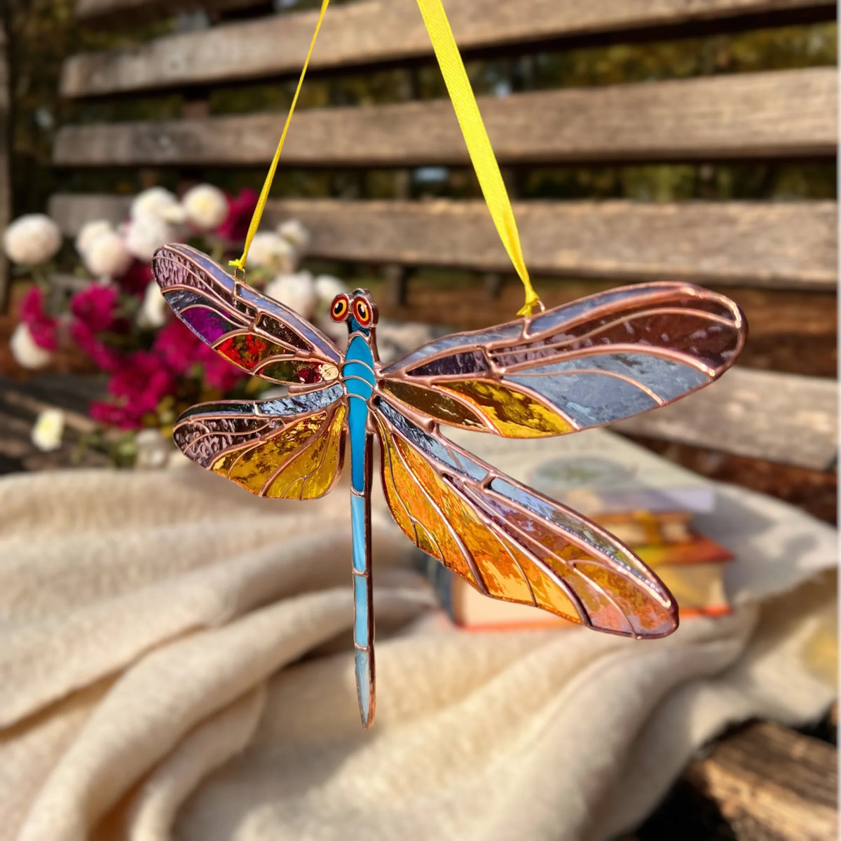 A stained glass dragonfly ornament with a bright blue body and amber, orange, and purple wings, hanging by a yellow ribbon in front of a wooden bench.