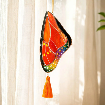 A stained glass butterfly suncatcher with a vibrant orange, red, green, blue, and purple wing design, hanging from a gold string with an orange tassel below, set against a soft-focus background of white curtains.