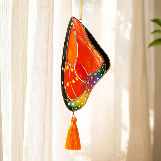 A stained glass butterfly suncatcher with a vibrant orange, red, green, blue, and purple wing design, hanging from a gold string with an orange tassel below, set against a soft-focus background of white curtains.