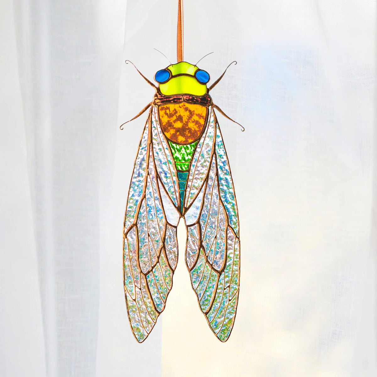 A stained glass cicada ornament hanging from a thin cord, featuring a green head and thorax, a bright yellow-orange abdomen, blue eyes, and translucent patterned wings against a soft, blurred green and pink background.