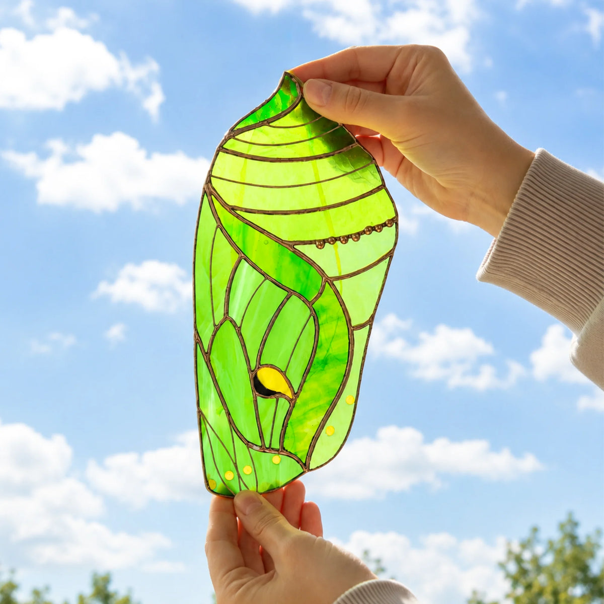 A stained glass suncatcher shaped like a luna moth chrysalis with a small yellow and black bird detail, held up by two hands against a bright blue sky with clouds.