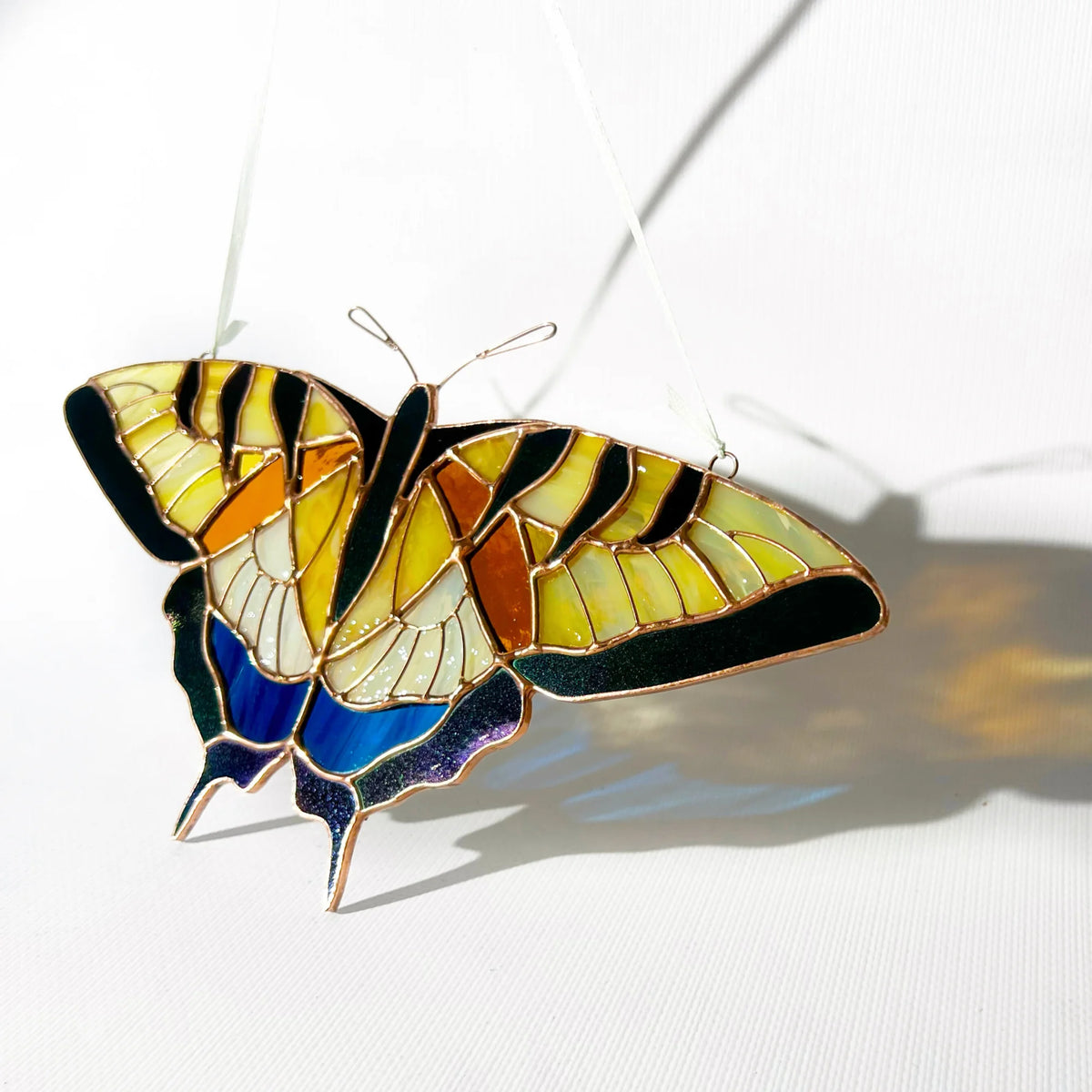 A stained glass swallowtail butterfly ornament with yellow, orange, black, blue, and white sections, suspended by thin wires against a white background with a soft shadow.