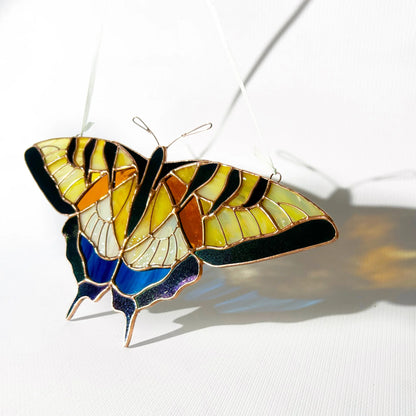 A stained glass swallowtail butterfly ornament with yellow, orange, black, blue, and white sections, suspended by thin wires against a white background with a soft shadow.