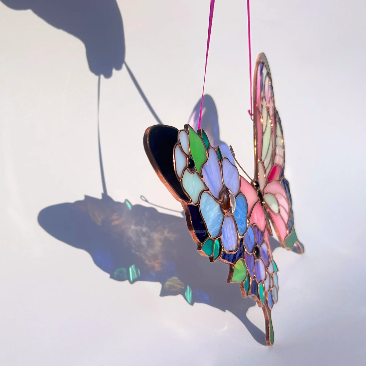 A stained glass butterfly ornament with a pink ribbon hanger, featuring multicolored glass panels in shades of blue, green, pink, and purple with copper-colored metal outlines, shown against a white background with a strong shadow.