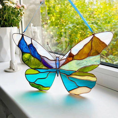 A stained-glass butterfly ornament with blue, green, yellow, white, and purple sections hangs on a white windowsill. A real window with outdoor greenery is visible in the background, along with candles and flowers.