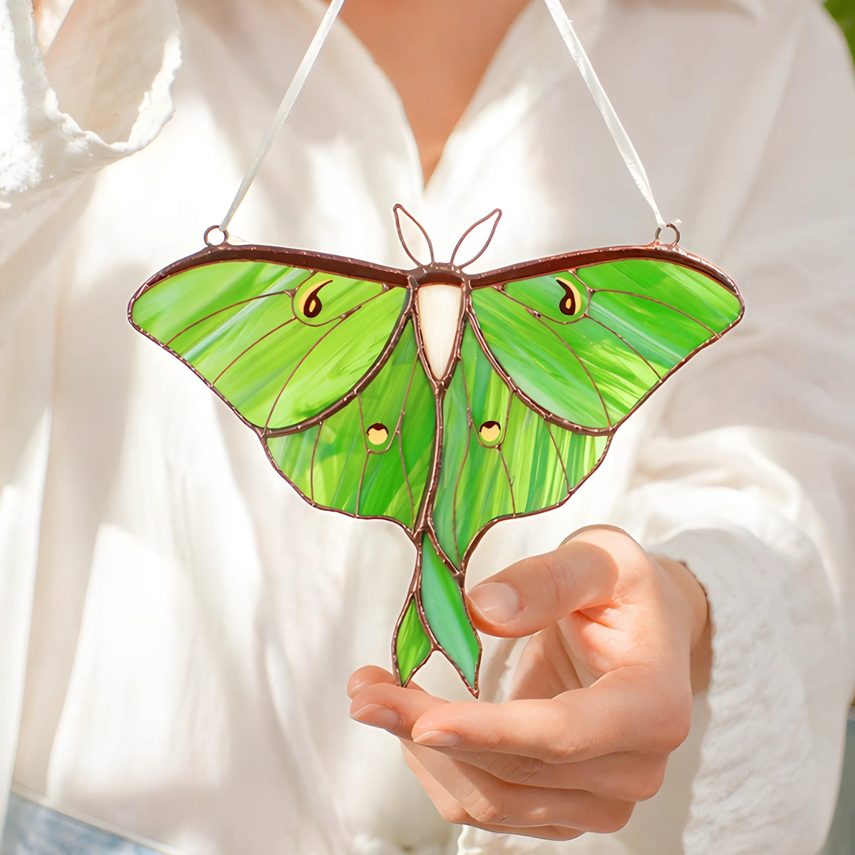 A green luna moth stained glass ornament with gold accents and white hanging strings, held by a person wearing a white shirt.