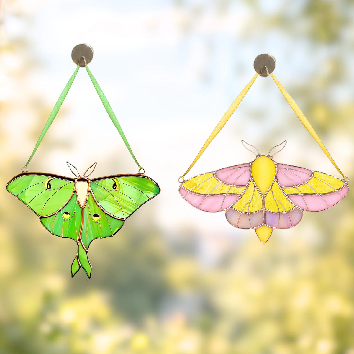 Two hanging stained-glass moth ornaments are shown against a soft blurred outdoor background. The left ornament is green with black outlines and small eye-like details, and the right ornament is pink and yellow with black outlines.