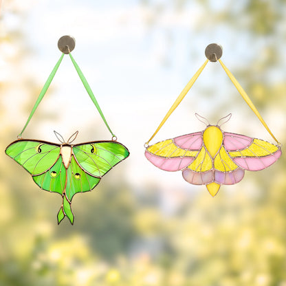 Two hanging stained-glass moth ornaments are shown against a soft blurred outdoor background. The left ornament is green with black outlines and small eye-like details, and the right ornament is pink and yellow with black outlines.