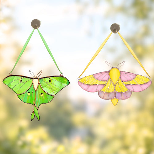 Two hanging stained-glass moth ornaments are shown against a soft blurred outdoor background. The left ornament is green with black outlines and small eye-like details, and the right ornament is pink and yellow with black outlines.