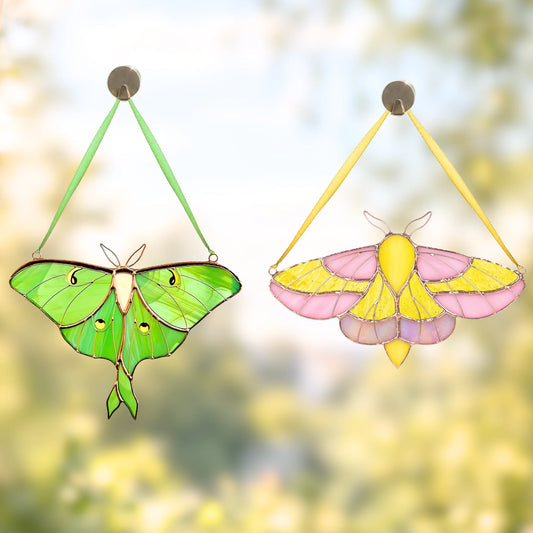 Two hanging stained-glass moth ornaments are shown against a soft blurred outdoor background. The left ornament is green with black outlines and small eye-like details, and the right ornament is pink and yellow with black outlines.