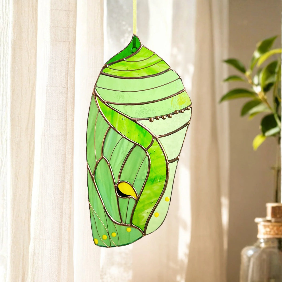 A stained glass luna moth chrysalis hanging ornament in shades of green with yellow accents, suspended in front of a window with sheer curtains.