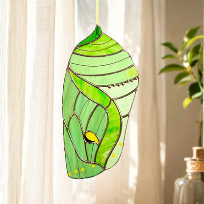 A stained glass luna moth chrysalis hanging ornament in shades of green with yellow accents, suspended in front of a window with sheer curtains.