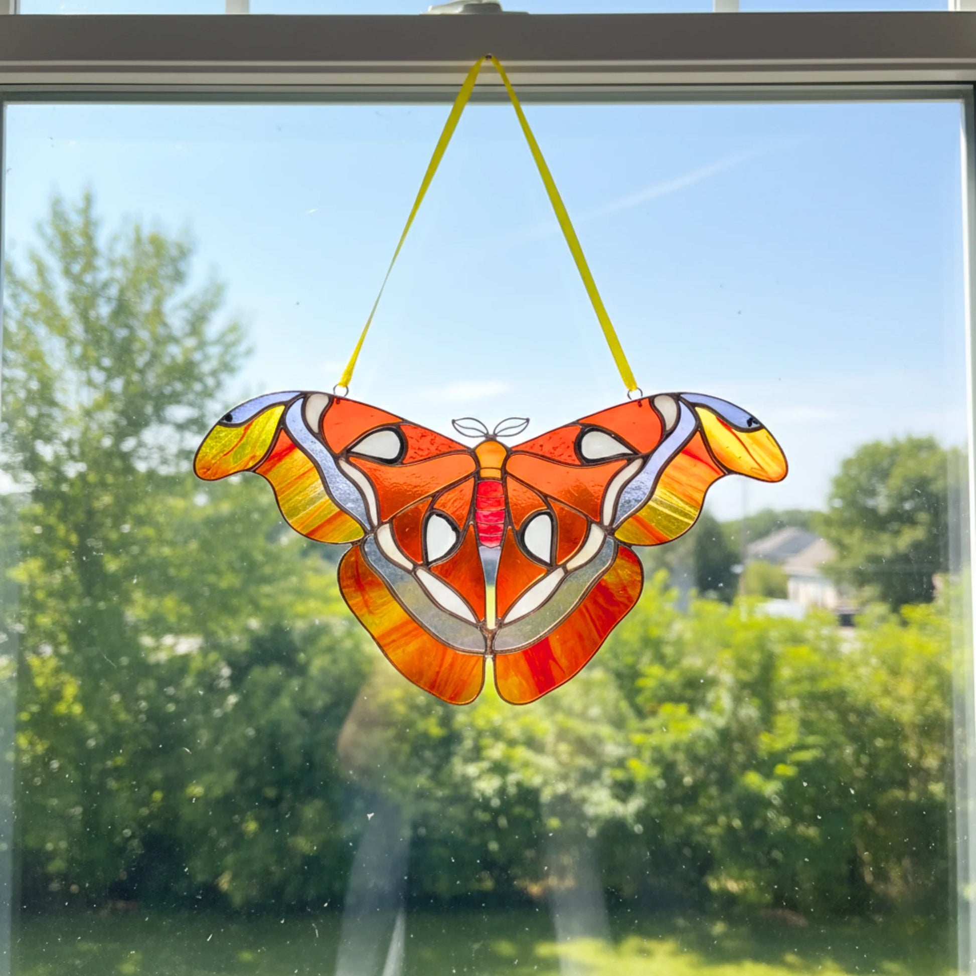 A stained-glass-style butterfly moth ornament with orange, red, yellow, white, and blue patterned wings hangs from a yellow ribbon in front of a window, with a blurred outdoor garden background visible.